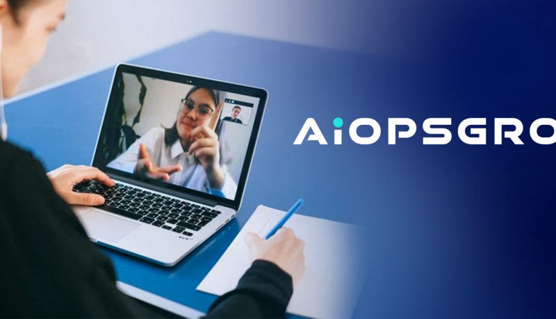 Una donna è impegnata in una videochiamata su un laptop, prendendo appunti su un blocco note accanto a sé. La donna sullo schermo sta usando il linguaggio dei segni, sottolineando l'accessibilità della comunicazione. L'ambientazione è professionale e il logo AIOPSGROUP è ben visibile sul lato destro dell'immagine, evidenziando l'attenzione dell'azienda verso soluzioni e tecnologie di comunicazione inclusive.