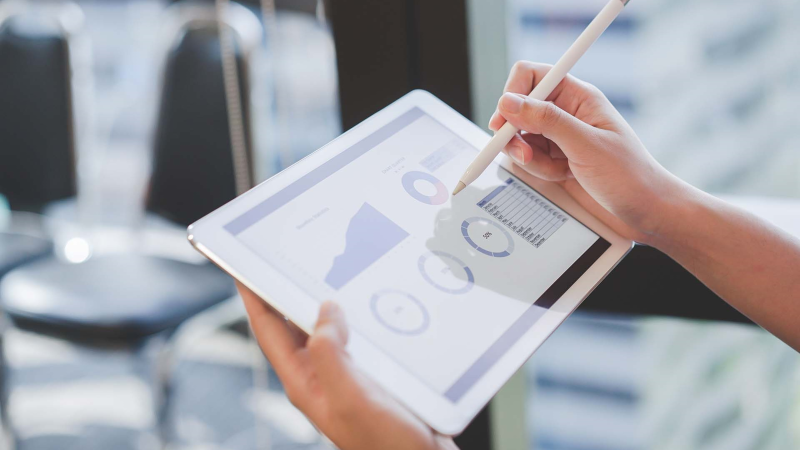 Närbild på en persons händer som håller en surfplatta och använder en stylus för att interagera med en digital analyspanel som visas på skärmen. Panelen visar olika diagram och grafer relaterade till företagets resultat. Bakgrunden är mjukt suddig och visar en ljus kontorsmiljö med tomma stolar, vilket antyder en miljö för professionell analys eller granskning.