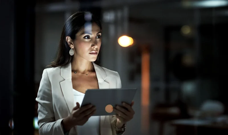 Een professionele vrouw in een lichtbeige pak, die aandachtig naar een tablet kijkt die ze vasthoudt. Haar geconcentreerde blik en de manier waarop ze in haar apparaat verdiept is, suggereren dat ze volledig in haar werk opgaat. De setting lijkt een kantoor 's avonds te zijn, met gedempte verlichting en een wazige achtergrond die bijdraagt aan de sfeer van late uren die aan belangrijke taken worden besteed. De verfijnde oorbellen en delicate ketting van de vrouw voegen een vleugje elegantie toe en benadrukken haar zelfverzekerde en beheerste houding in een zakelijke omgeving.