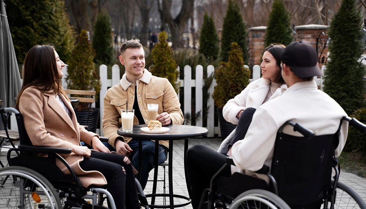 Quatre amis, dont deux en fauteuil roulant, profitent d'une réunion informelle en plein air dans un café. Ils sont assis autour d'une table avec des boissons et des collations, discutent et sourient. Le cadre est chaleureux, avec une clôture blanche et de la verdure en arrière-plan, créant une atmosphère accueillante et inclusive.