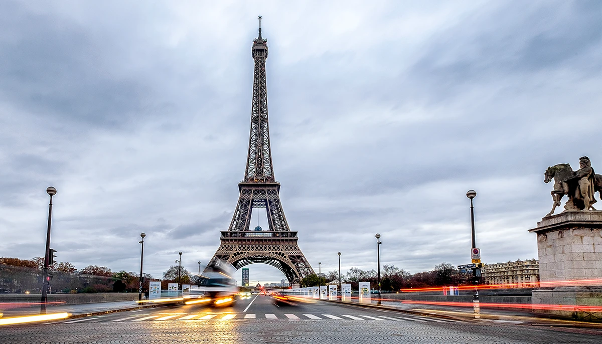 La tour Eiffel à Paris, en France, se dresse sous un ciel nuageux, avec les lumières floues des véhicules qui passent dans la rue au premier plan, créant un contraste dynamique entre le mouvement et ce monument immobile.