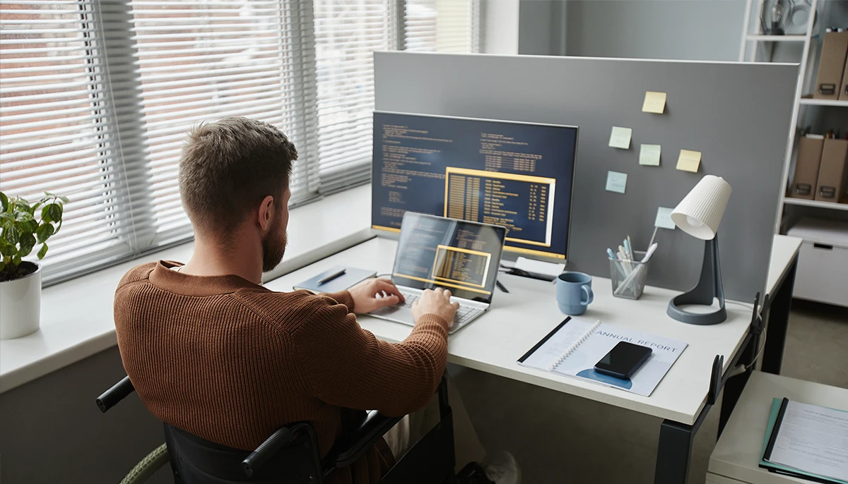 Un homme en fauteuil roulant travaille à un bureau avec un ordinateur portable et un écran externe affichant du code, effectuant un audit manuel d'accessibilité numérique dans un environnement de bureau moderne.