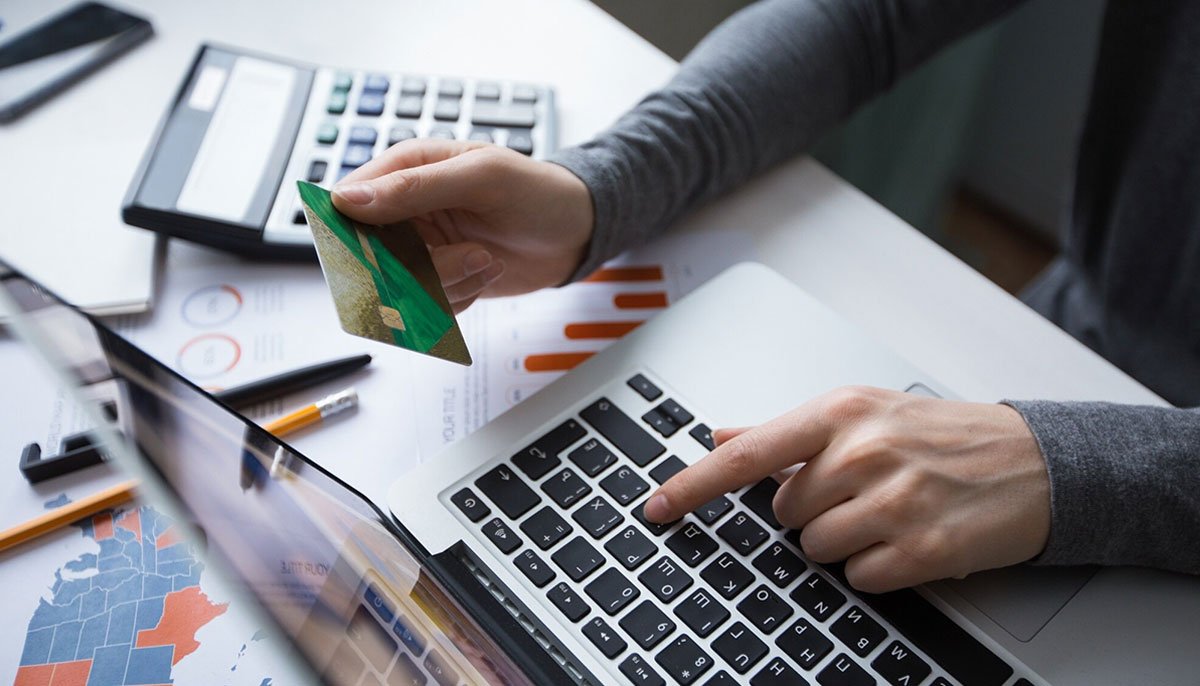 Gros plan sur une personne effectuant un paiement en ligne, tenant une carte de crédit verte dans une main et tapant sur un ordinateur portable de l'autre. Des documents financiers, une calculatrice et un stylo sont visibles sur le bureau, indiquant une activité liée à la gestion budgétaire ou au commerce électronique.