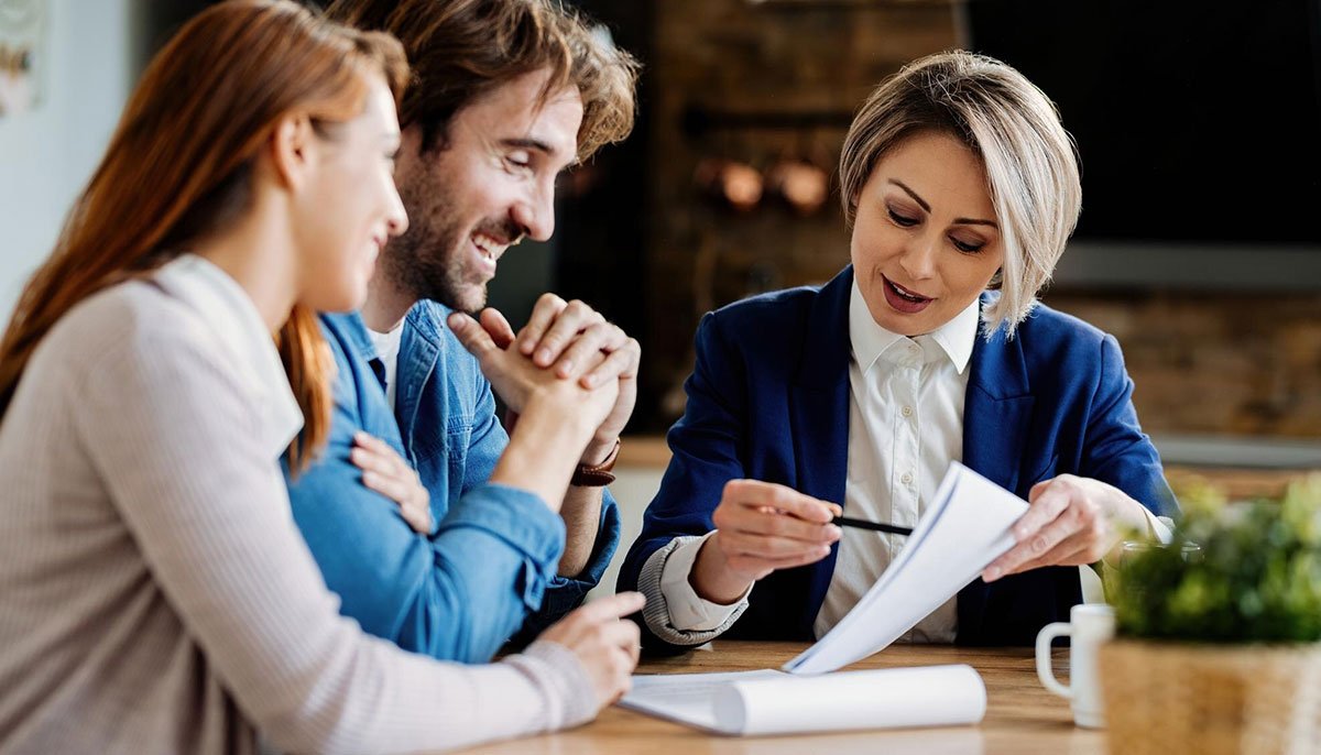 Un conseiller financier ou un consultant montre des documents à un couple souriant lors d'une réunion. Le couple semble engagé et positif alors qu'il examine ensemble des documents à une table dans un bureau ou un cadre domestique bien éclairé.