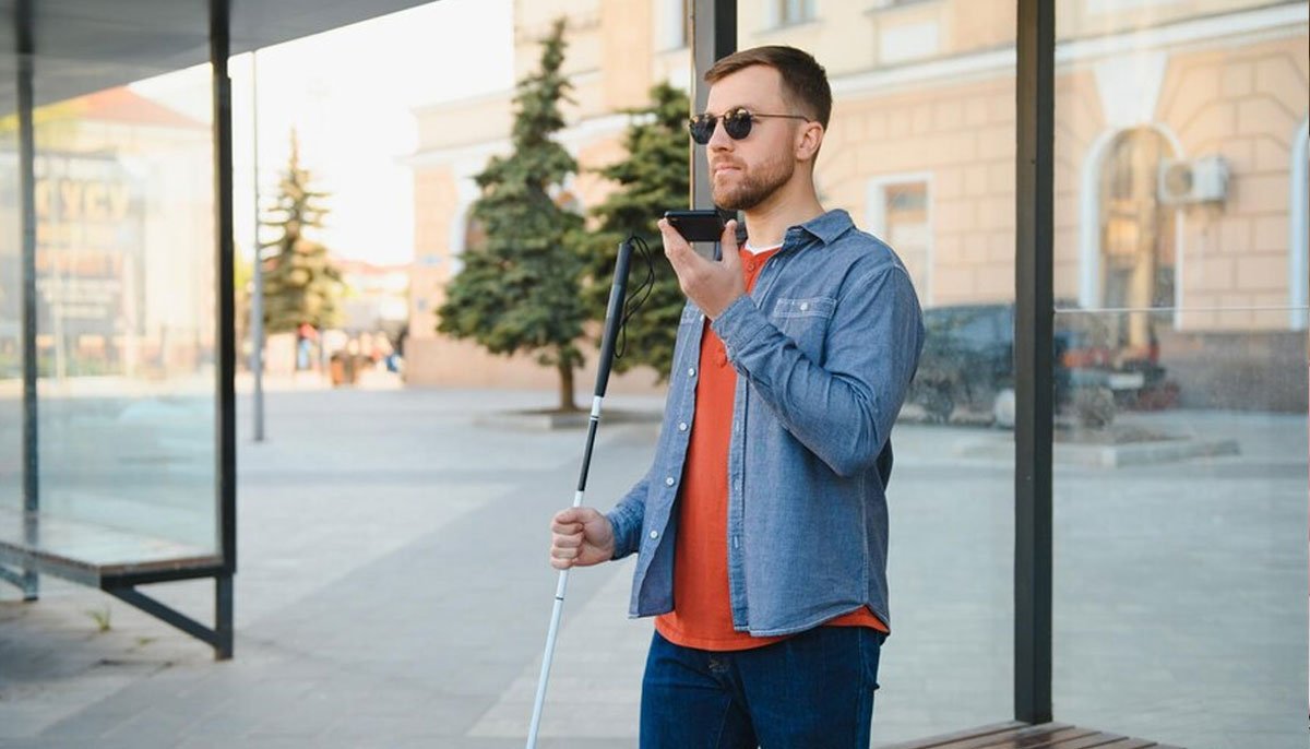 Un homme aveugle portant des lunettes de soleil se tient à l'extérieur près d'un abri en verre, tenant une canne blanche dans une main et parlant dans un smartphone de l'autre. Il semble utiliser un assistant vocal ou une application de navigation dans un environnement urbain.