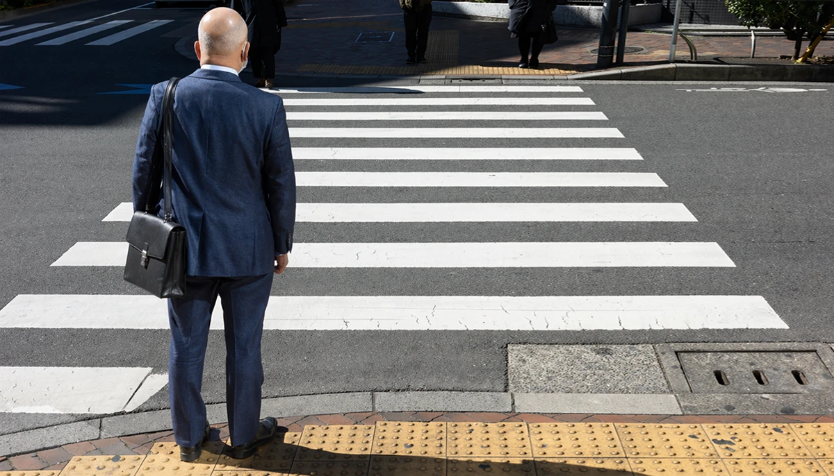 Een man in een blauw pak staat aan de rand van een zebrapad, met zijn gezicht naar de straat gericht, met braillebestrating onder zijn voeten en andere voetgangers in de verte.