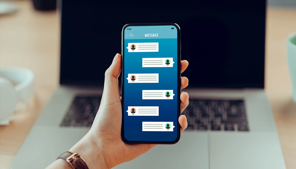 Une main tenant un smartphone sur lequel s'affiche une application de messagerie avec des conversations.